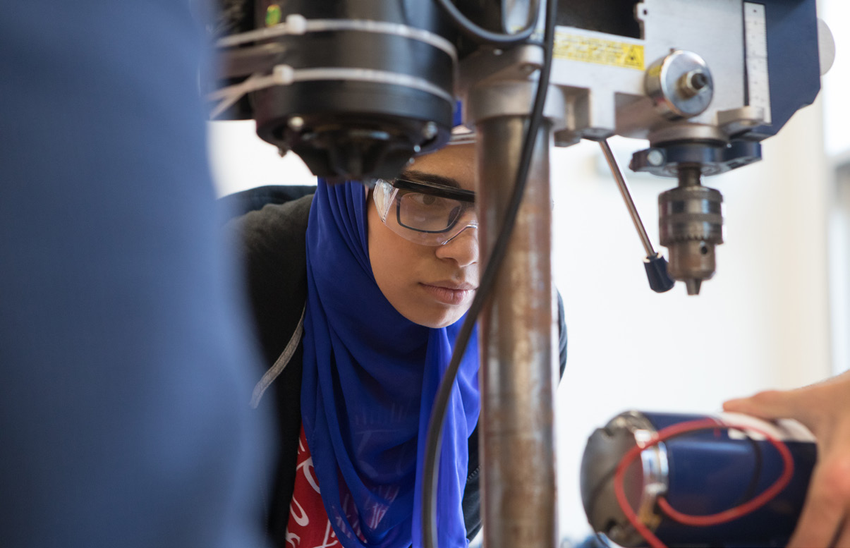 a student wearing goggles works with a drill press